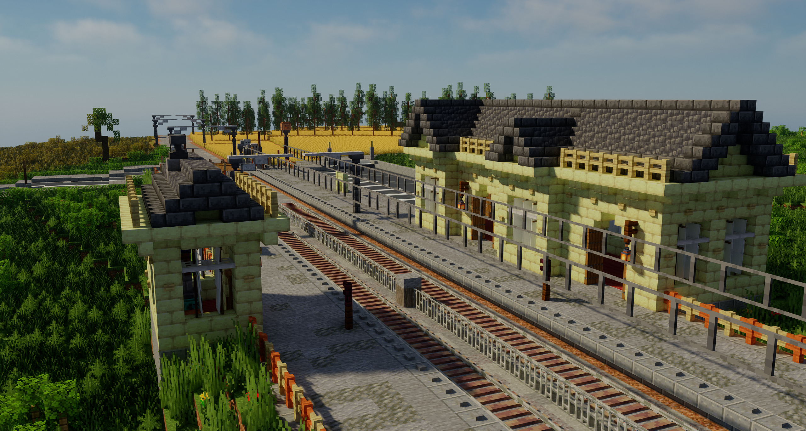 An image of St. Charles-on-the-Sea Station's platforms and station buildings. A railway crossing, wheat fields and vegetation are visible in the background.